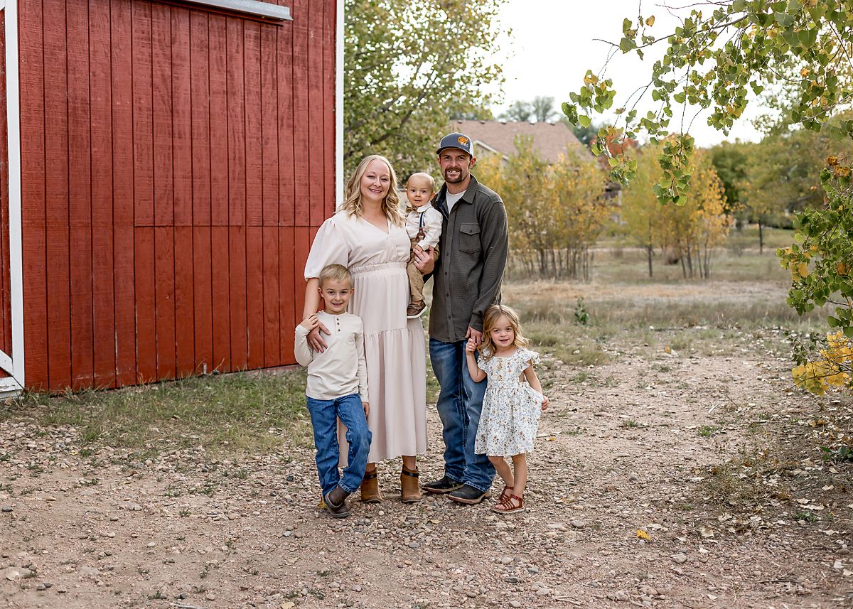 Family of five posing outdoors near a red barn with fall foliage in background.