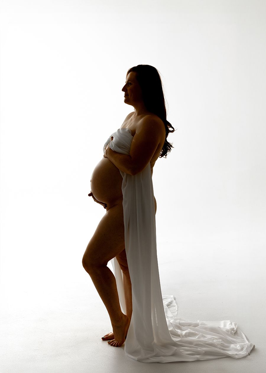 Pregnant woman in white crop top and flowing white skirt poses sideways in a bright studio maternity photo.