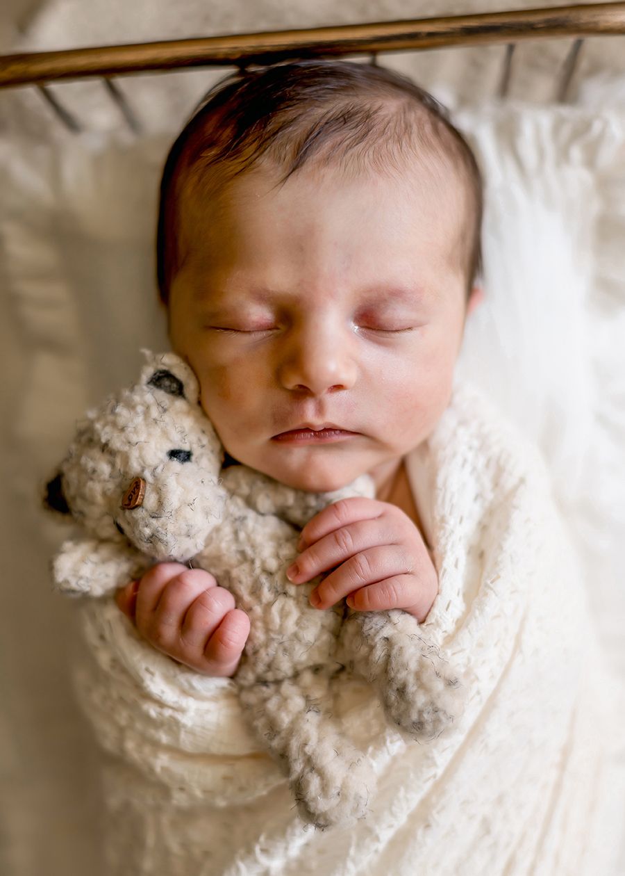 Sleeping newborn baby swaddled in cream wrap, holding a small teddy bear, peaceful nursery portrait.