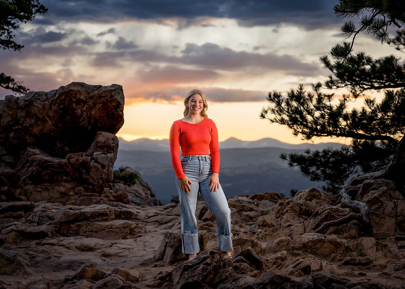 Teen girl in red top stands on rocky mountain summit at dramatic sunset.