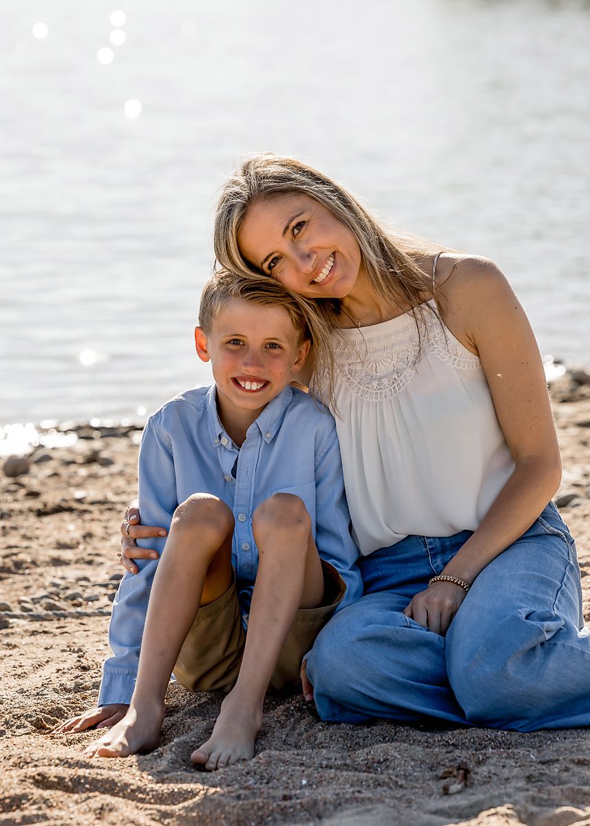 A mother and young son sit together smiling on a sandy lakeside beach.