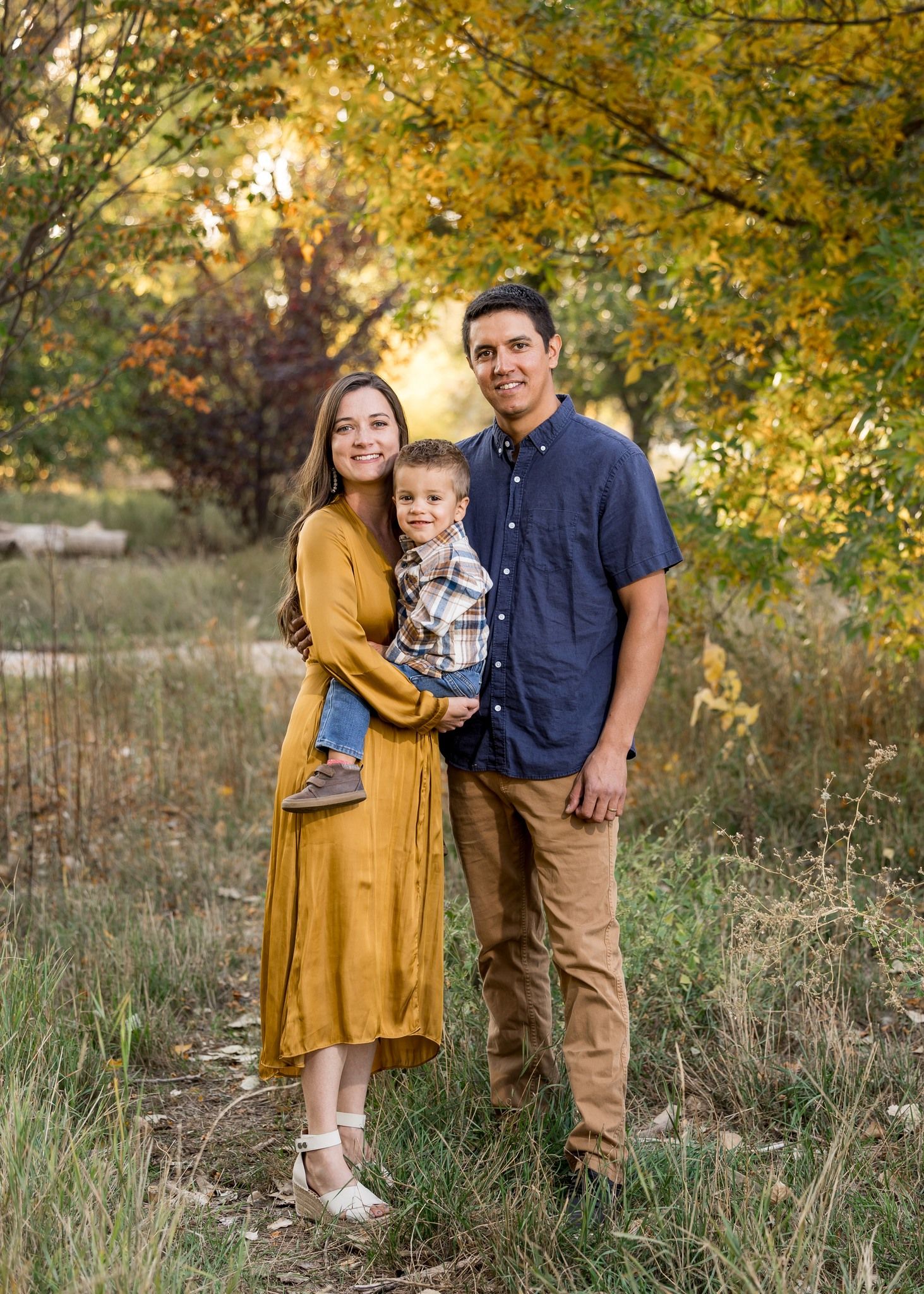 Family of three posing in a golden autumn meadow, mother in mustard dress holding toddler.