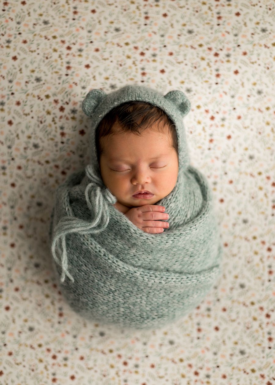 Sleeping newborn baby wrapped in a sage green knit swaddle with bear ear bonnet on a floral background.