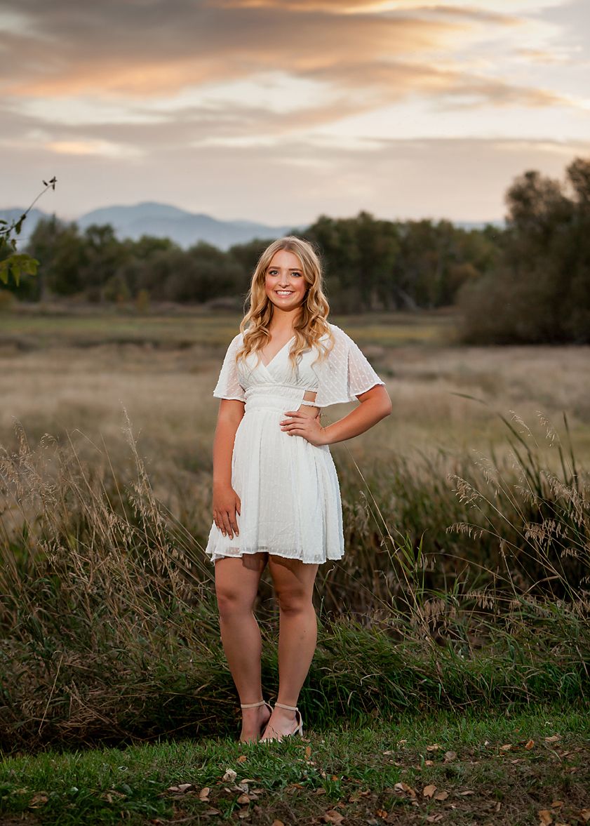 Young woman in white dress poses in golden field at sunset with mountain backdrop.