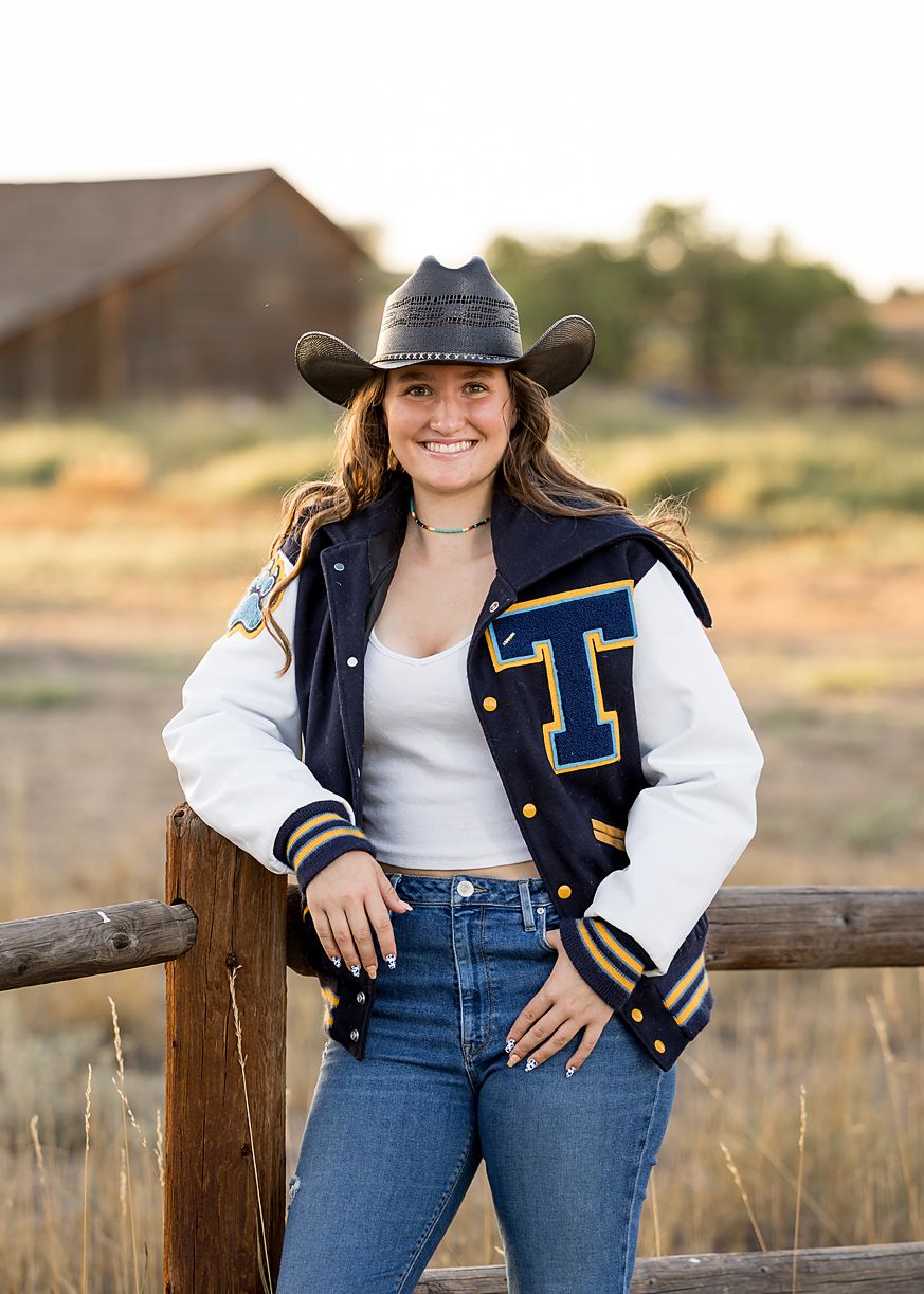 Smiling young woman wearing varsity jacket and cowboy hat leaning on wooden fence in rural field at sunset.
