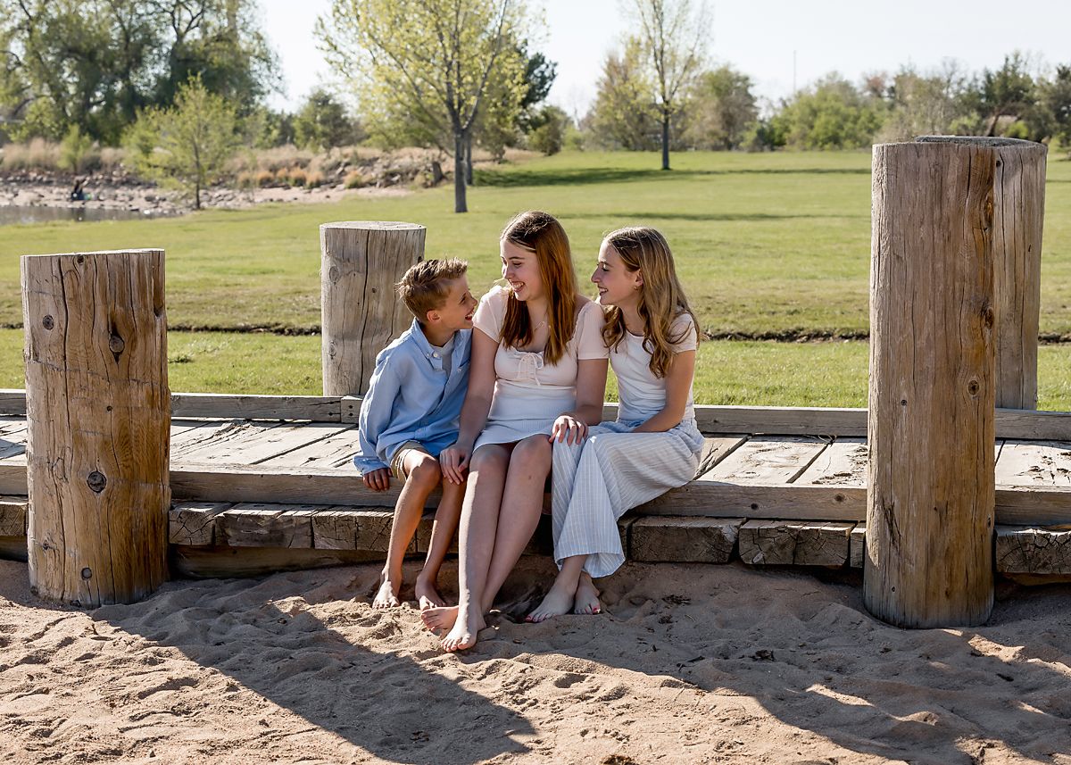 A mother sits with two children on a wooden dock near a grassy park.