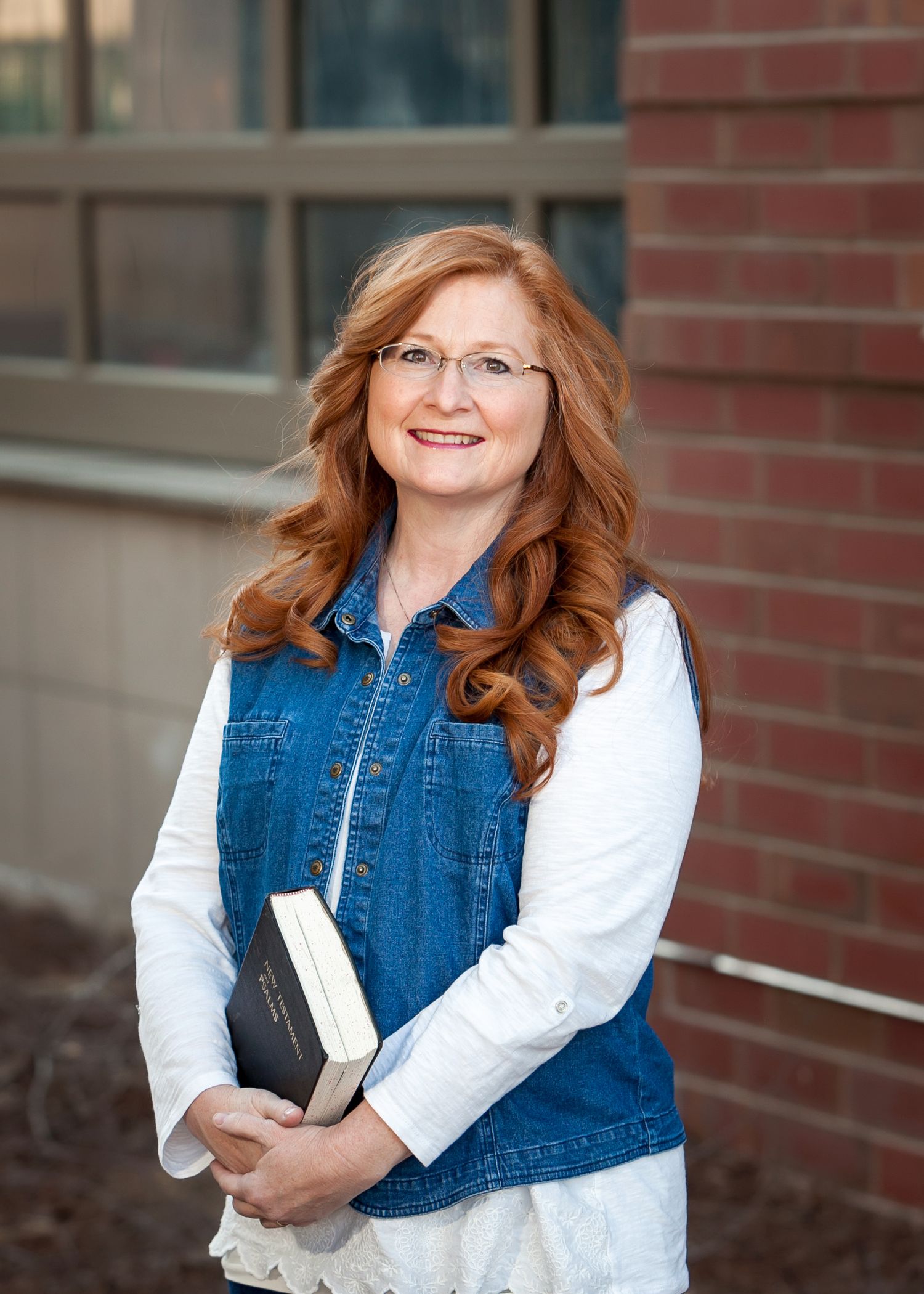 Smiling redhead woman holding a Bible outdoors near a brick wall in denim vest.
