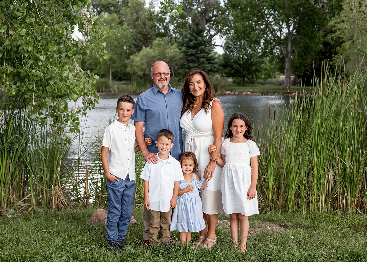 Family of six posing outdoors near a pond surrounded by lush green trees and grass on a sunny day.