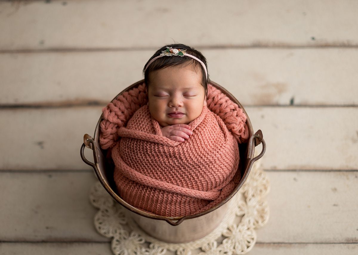 Newborn baby wrapped in pink knit swaddle with floral headband, sleeping peacefully in a copper bucket on white wood.