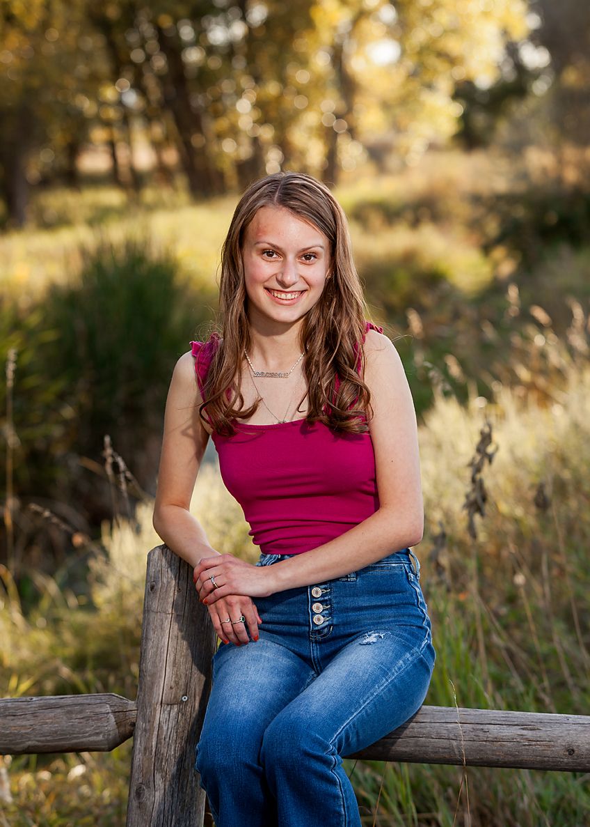 Smiling teenage girl in red top and jeans leans on rustic wooden fence for fall senior portrait.