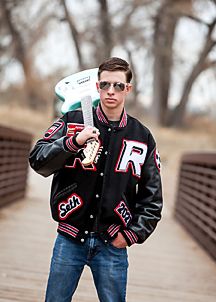 Young man in a varsity jacket holding a guitar on a wooden bridge wearing sunglasses outdoors.