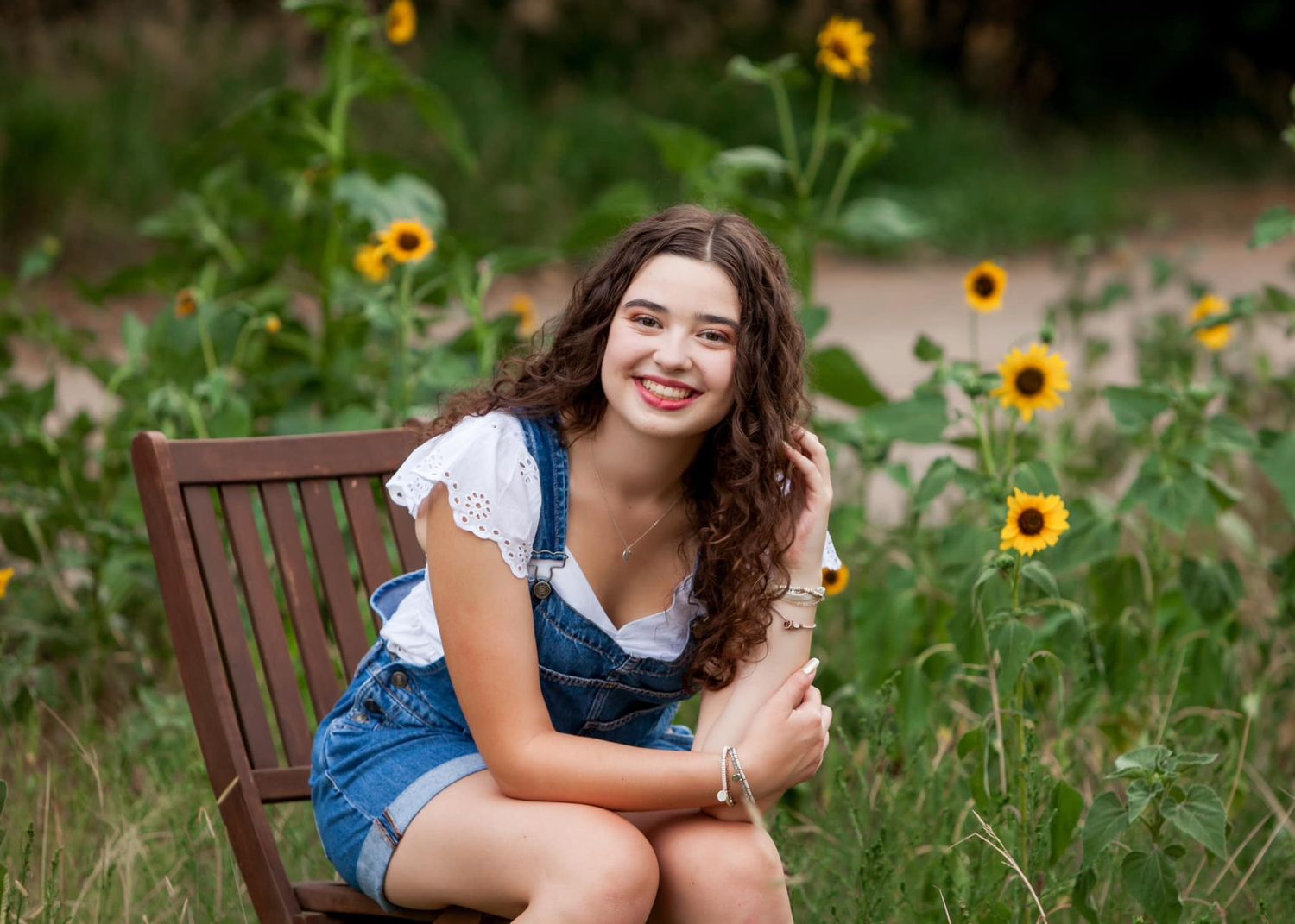 Smiling teenage girl sitting in a wooden chair surrounded by blooming sunflowers.