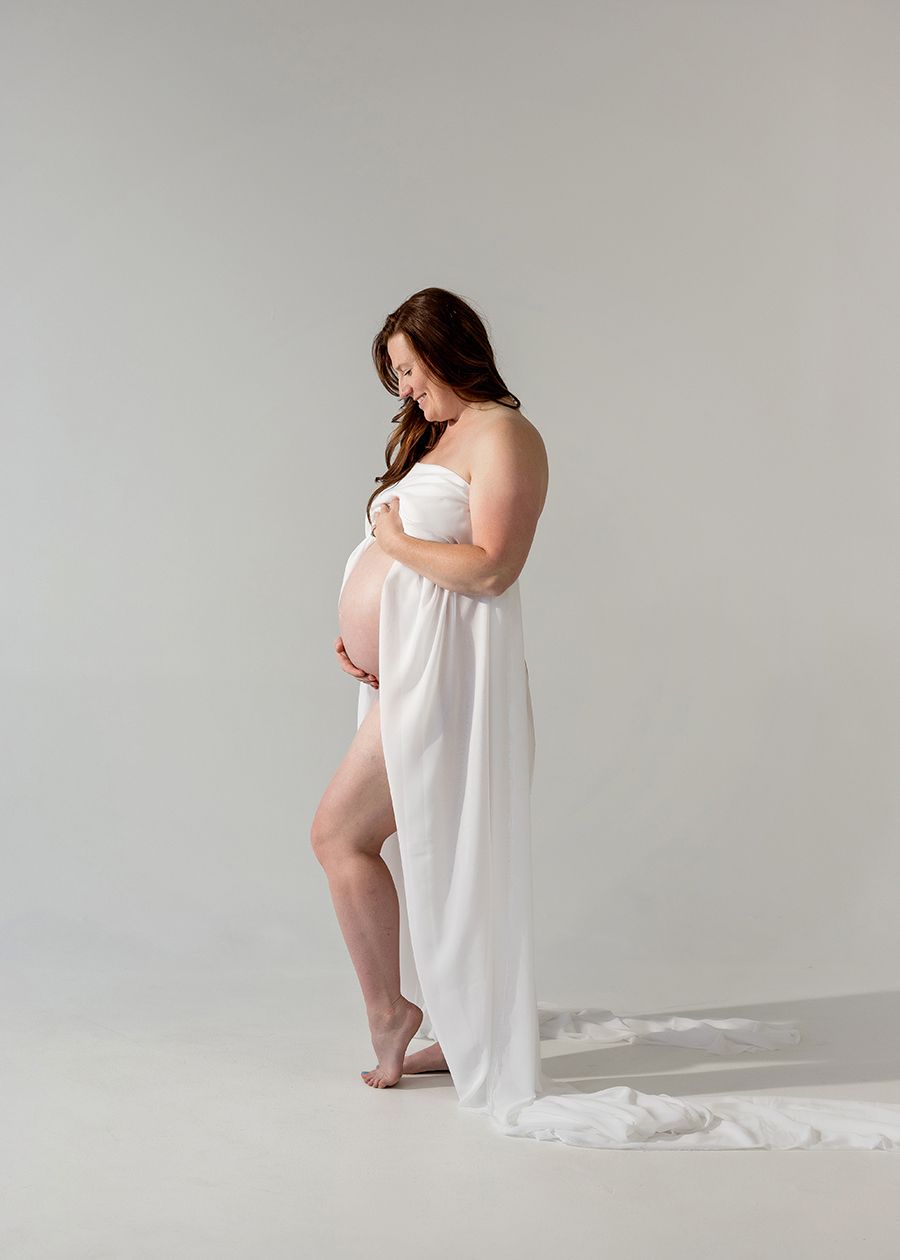 Pregnant woman in white draped fabric poses gracefully in a studio maternity portrait against a soft gray background.