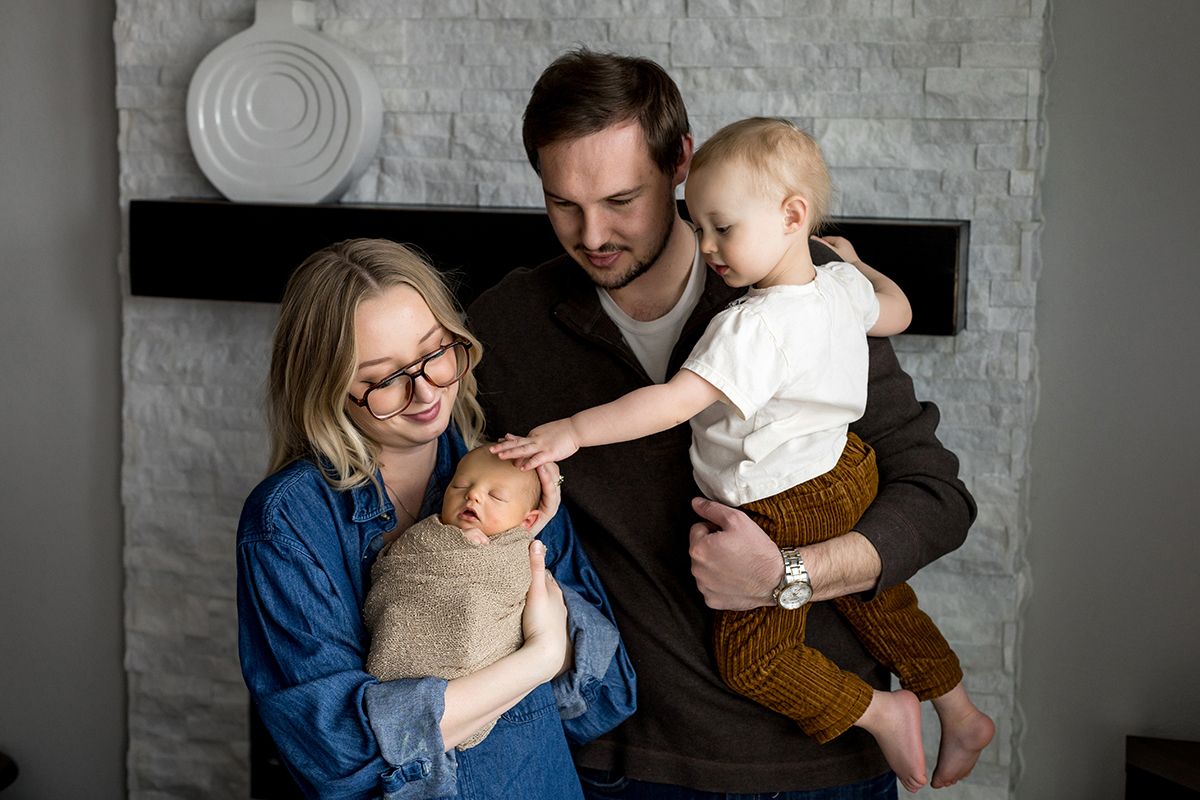 A young family poses warmly by a fireplace, with mom holding a swaddled newborn and dad carrying a toddler.
