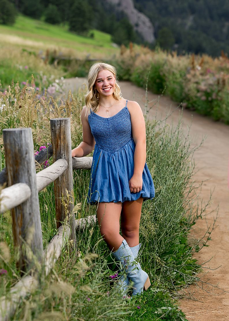 Blonde teen girl in blue denim dress and cowboy boots poses by fence on mountain trail.