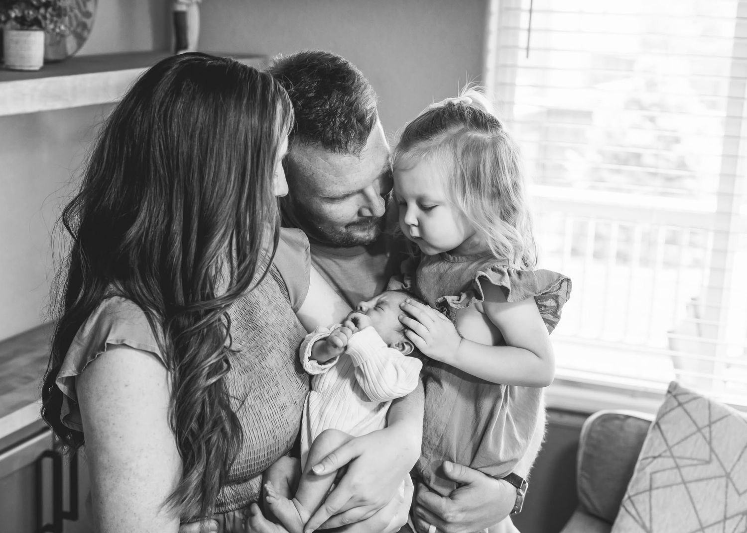 Black and white family portrait of parents and toddler girl lovingly gazing at newborn baby indoors.