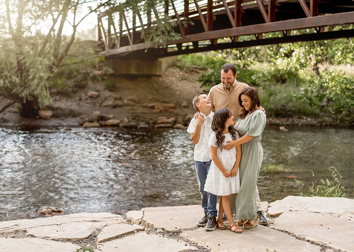 Happy family of four posing by a river with a rustic bridge backdrop during an outdoor photography session.