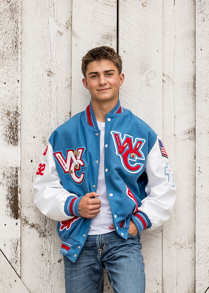 Teen boy in blue varsity letterman jacket poses against white weathered wood wall.