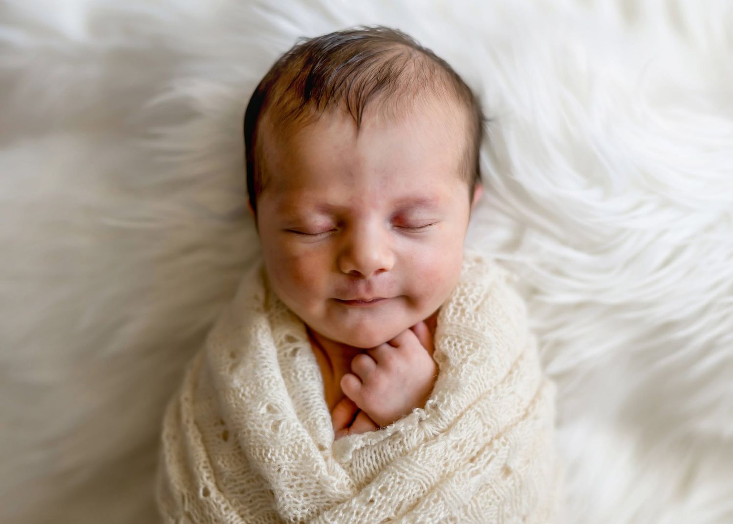 Peaceful newborn baby swaddled in a cream knit wrap smiling while resting on white fur.