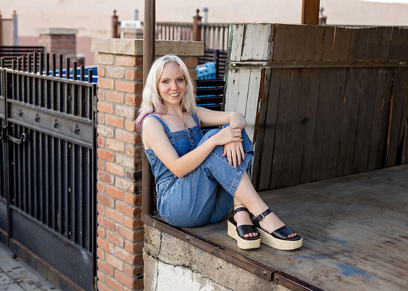 Young woman with blonde hair sitting on a brick ledge wearing a denim jumpsuit and platform sandals outdoors.