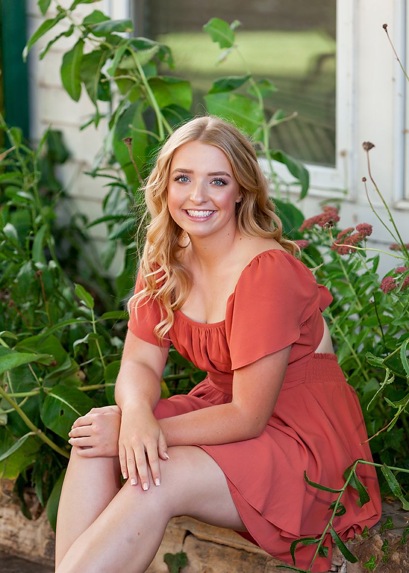 Young blonde woman in a coral off-shoulder dress sitting outdoors among lush green plants, smiling brightly.