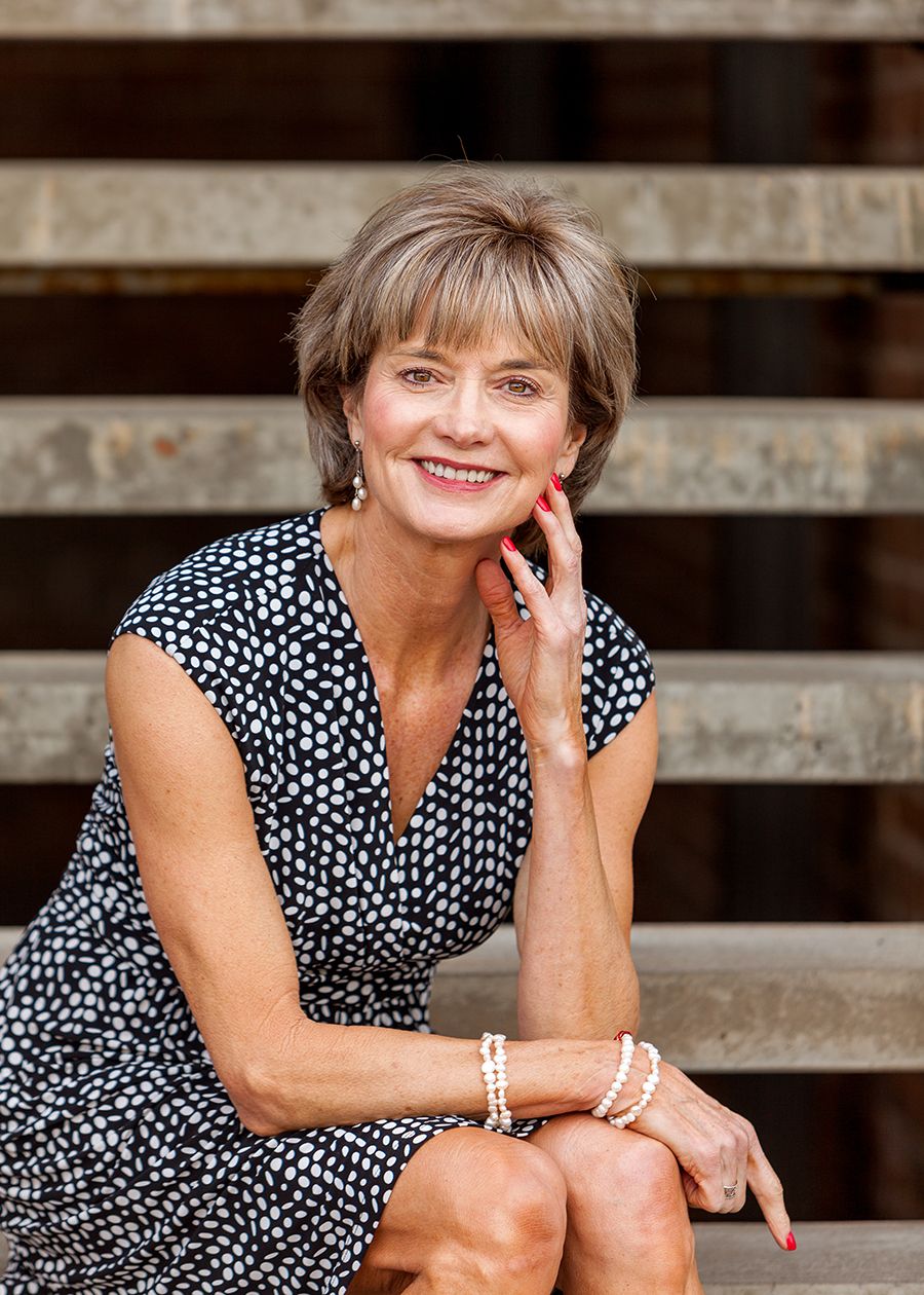 Smiling middle-aged woman in polka dot dress sitting on stone steps, wearing pearl jewelry, posing for a portrait photo.