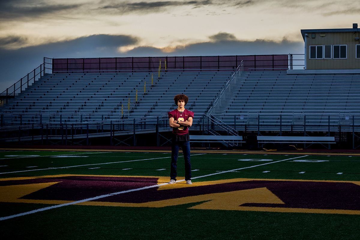 Young athlete holds football on stadium field at dusk, with bleachers and dramatic cloudy sky in background.