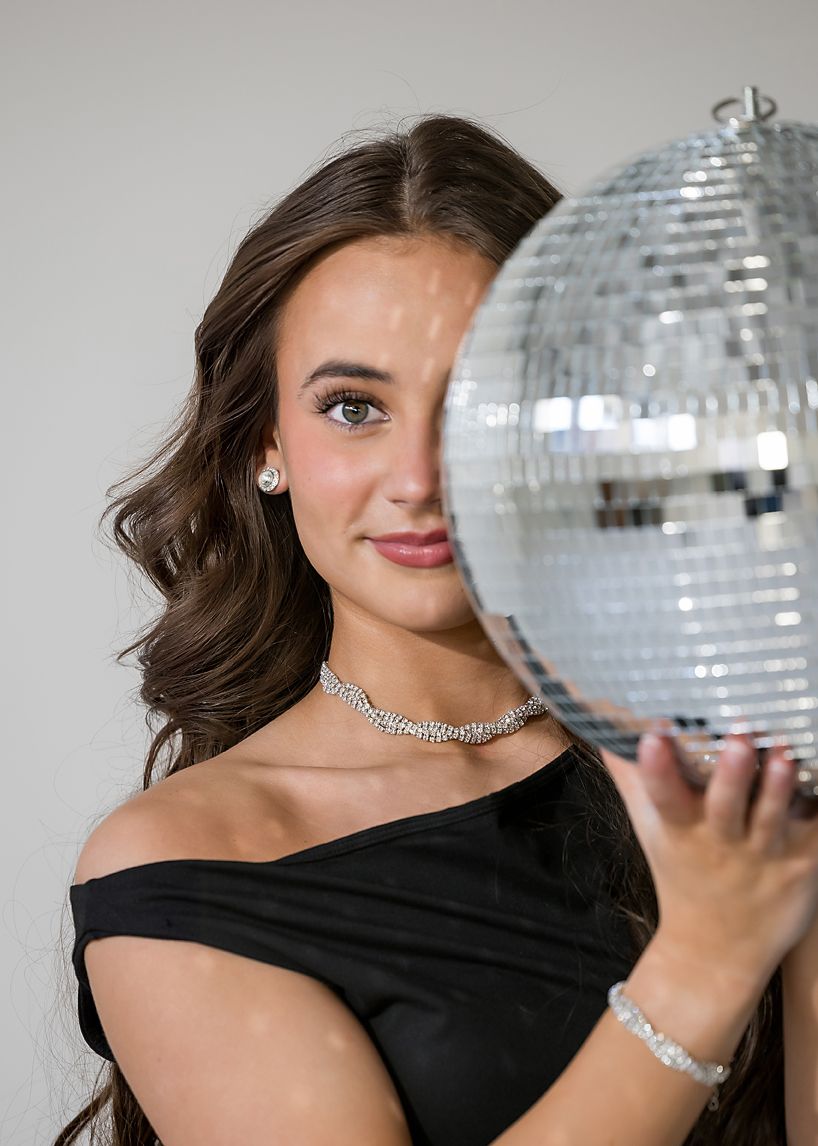Elegant brunette teen in black off-shoulder dress poses with a sparkling disco ball indoors.