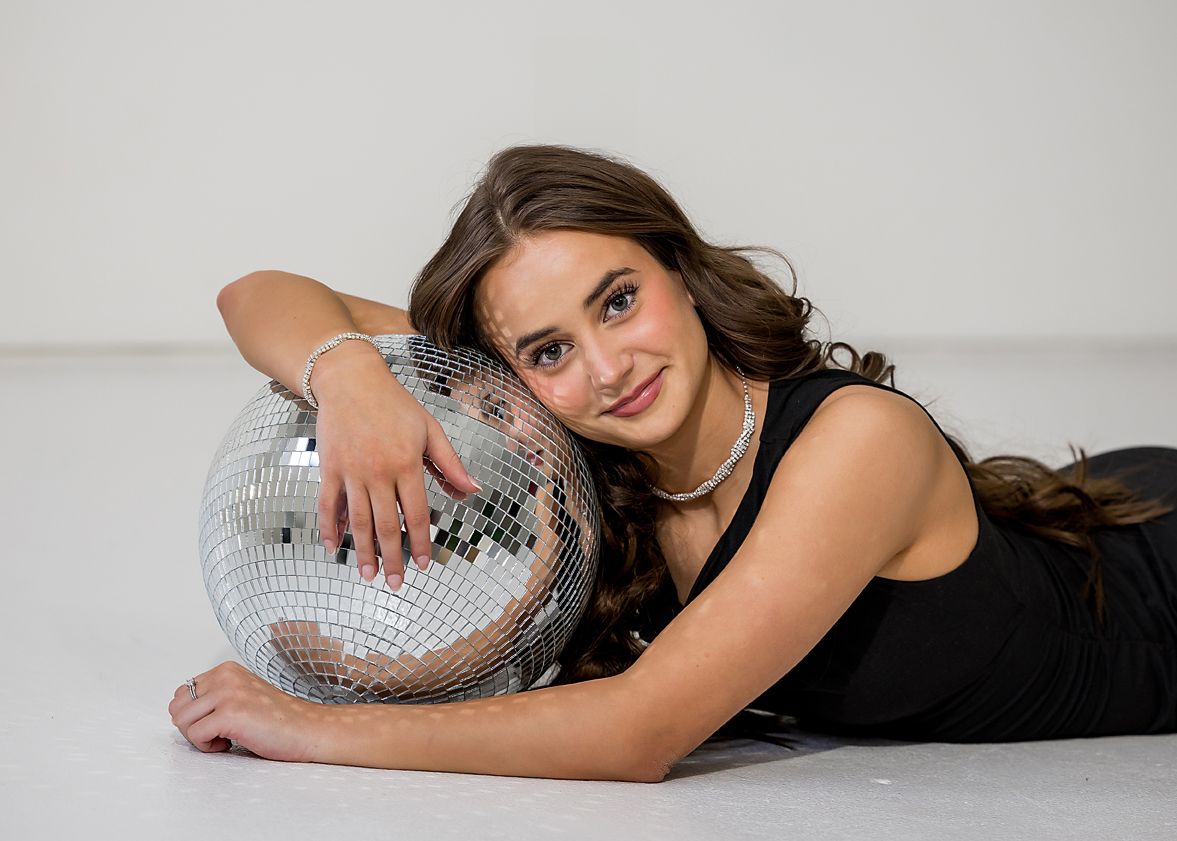 Young woman lying on floor smiling while leaning on a large mirrored disco ball.