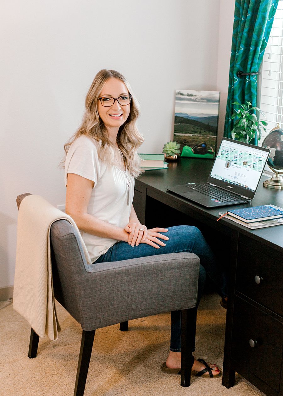 Smiling blonde woman with glasses sitting at a dark wood desk with a laptop in a bright home office.
