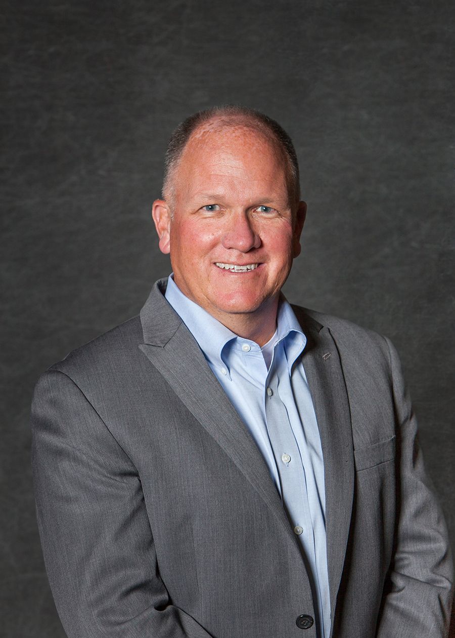 Professional headshot of a smiling middle-aged man in a gray blazer and light blue shirt against a dark background.