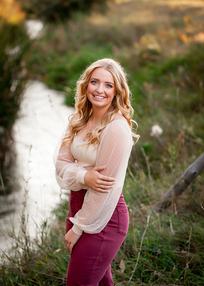 Smiling blonde teenage girl in pink pants and white blouse posing outdoors near a stream for senior photos.