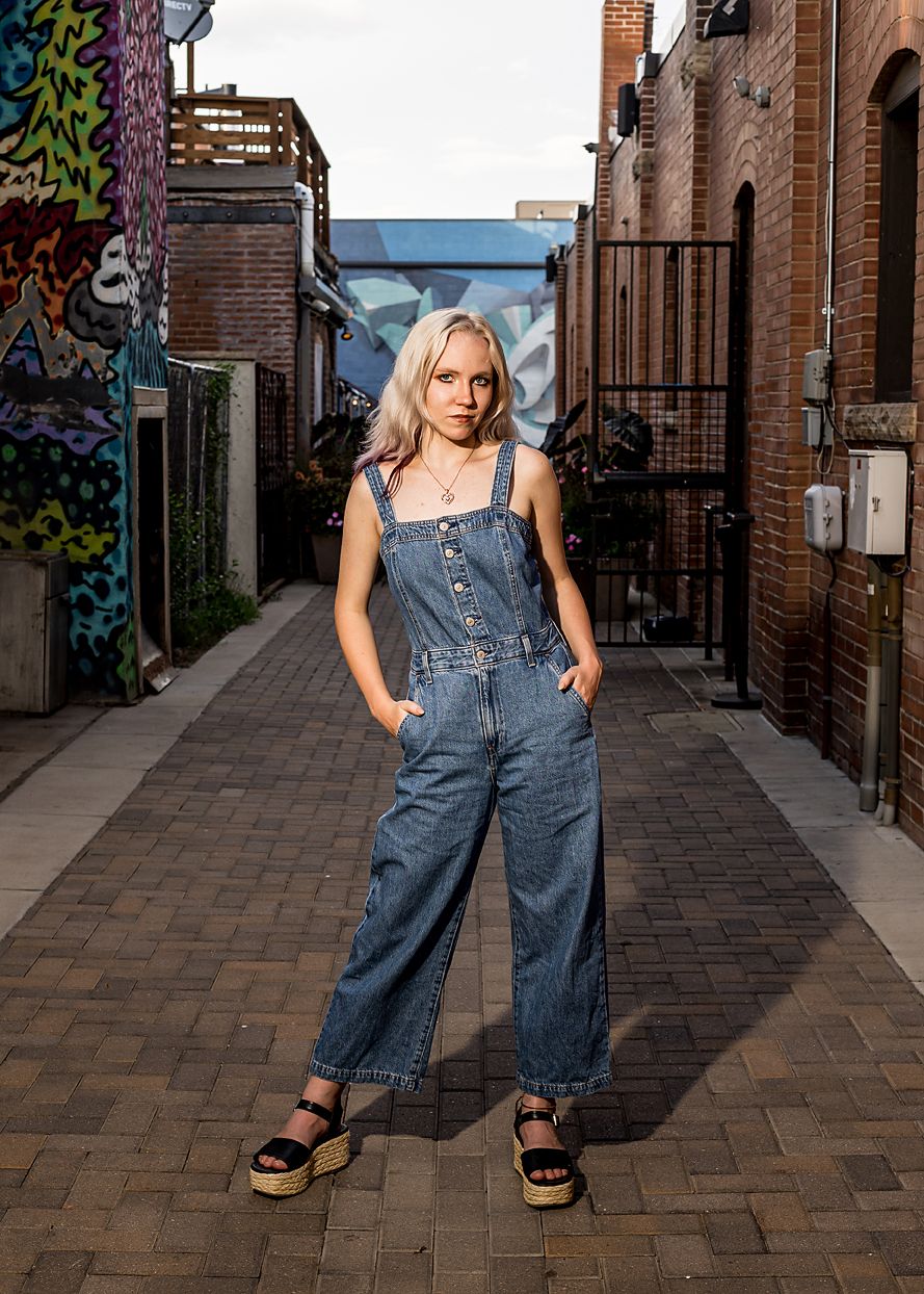 Young woman in denim overalls and platform sandals poses confidently in an urban alley with colorful murals.