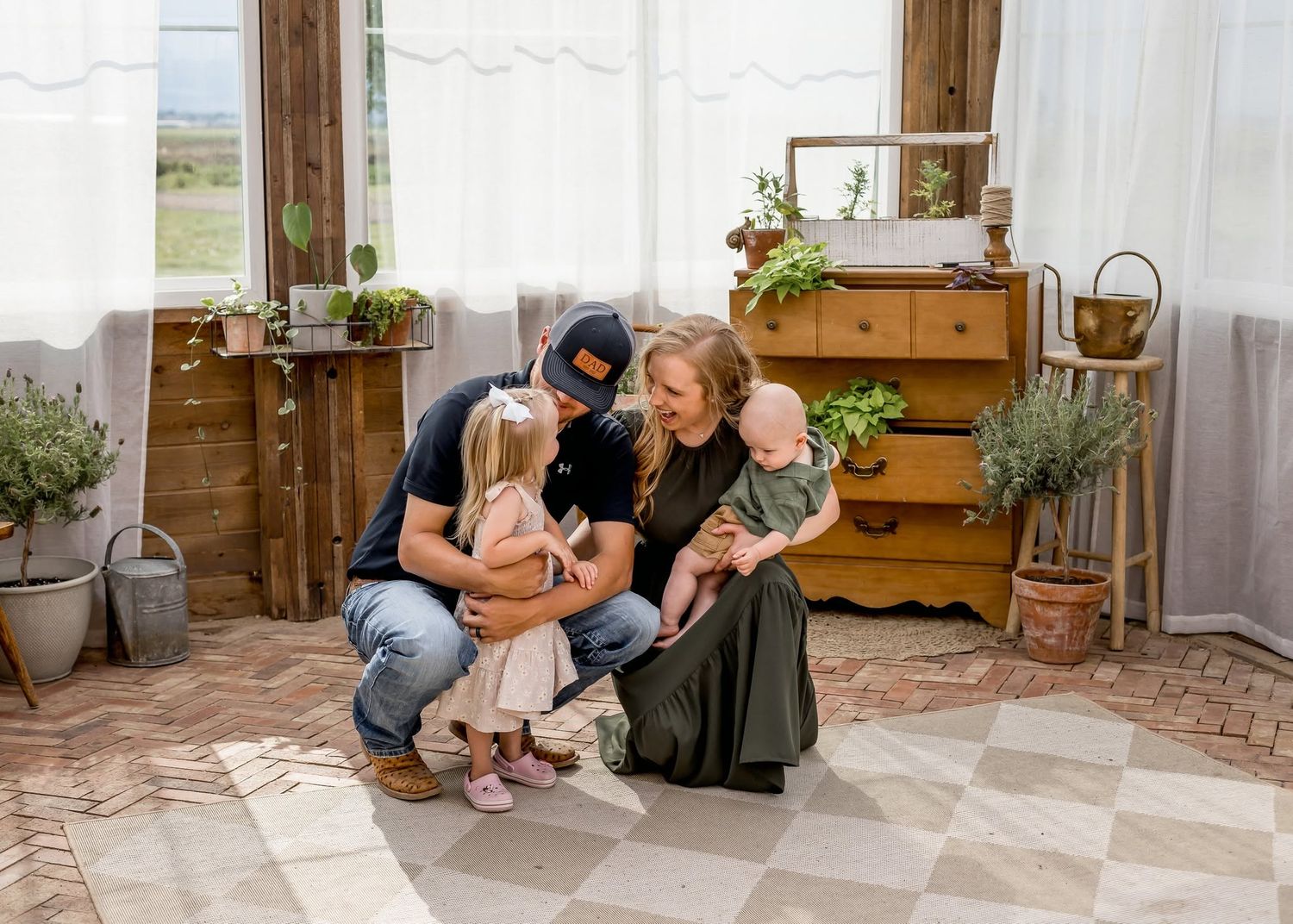 Family of four gathered together looking at a book in a rustic decorated room with wooden dresser and plants.