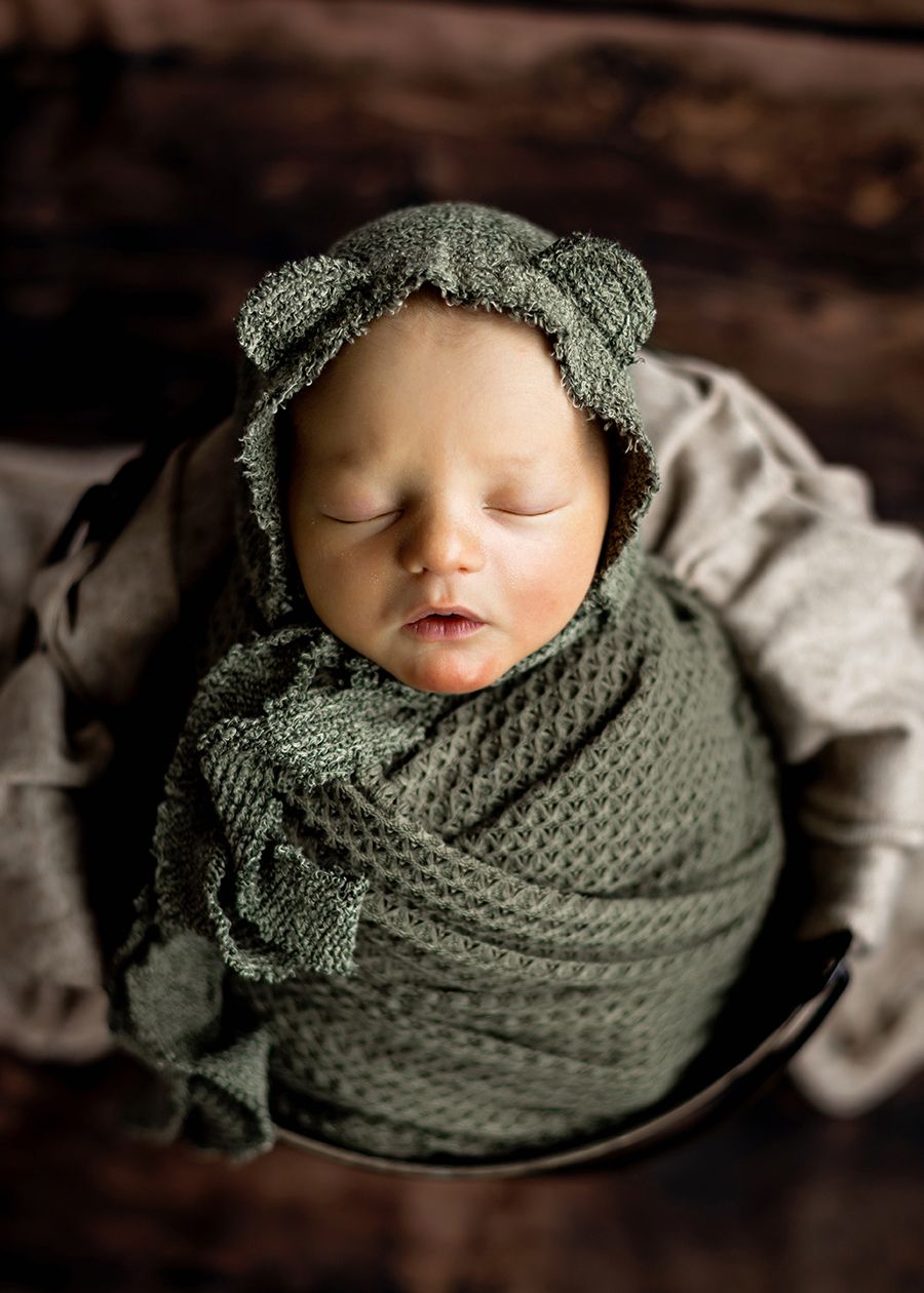 Sleeping newborn wrapped in green knit swaddle and bear ear bonnet inside a wooden bowl.