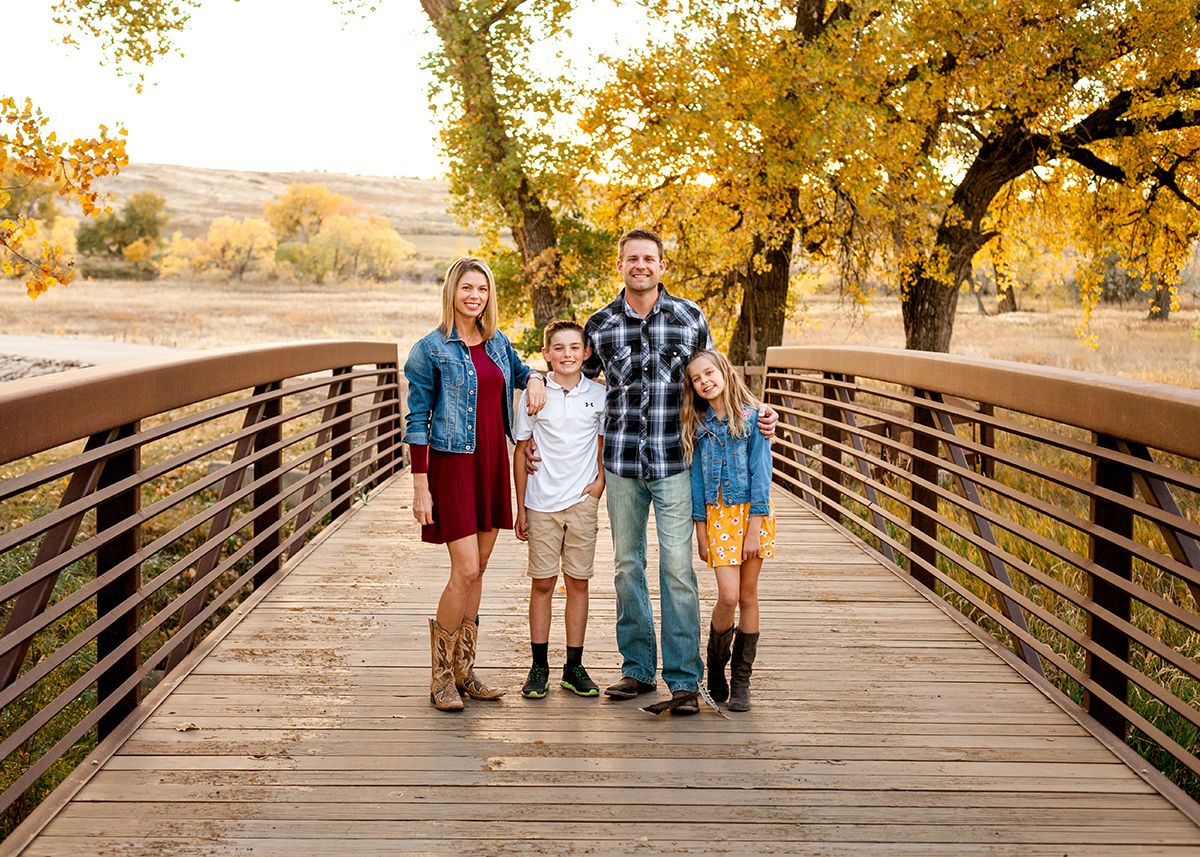 Family of four smiling on a wooden bridge surrounded by golden autumn trees.