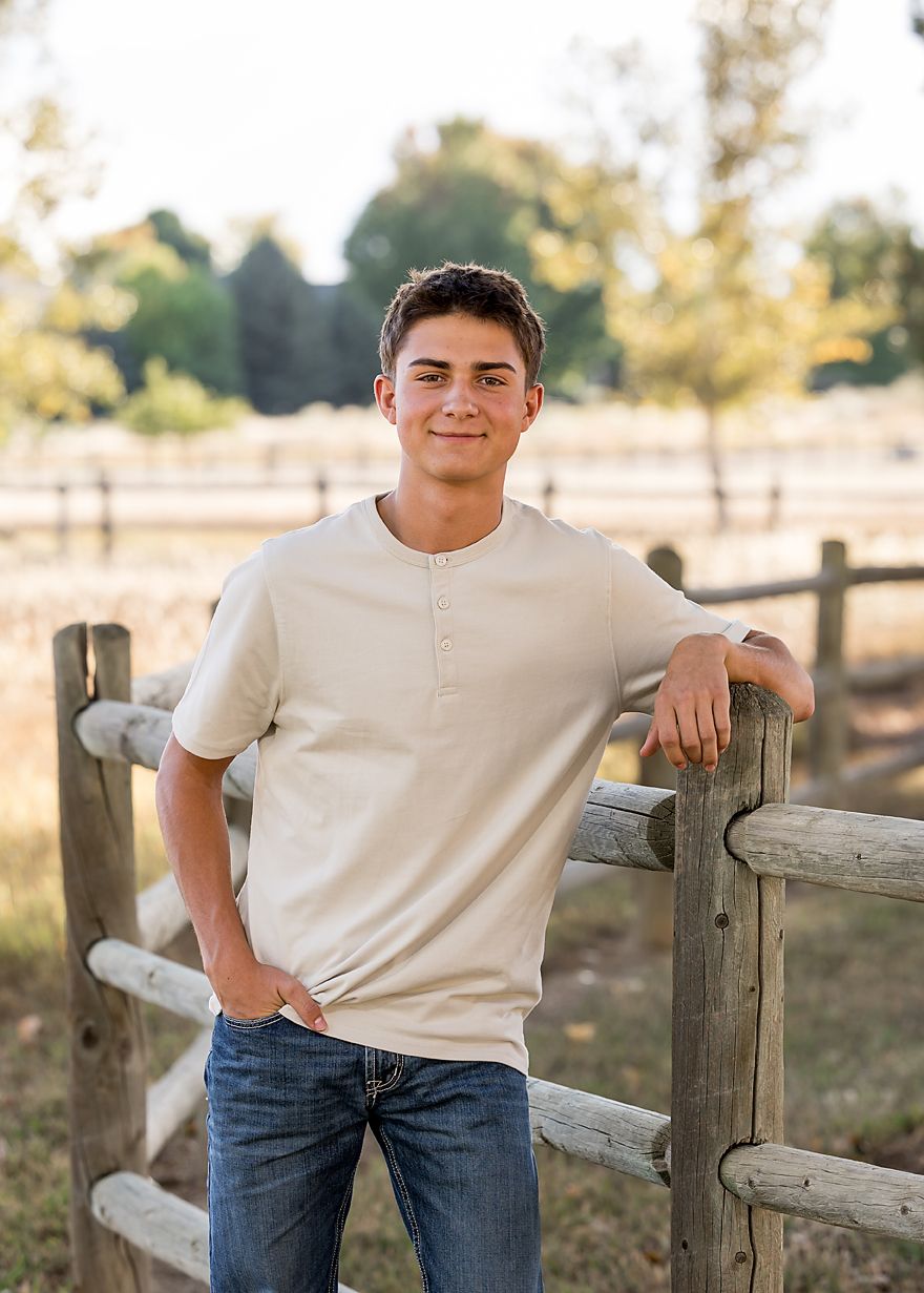 Teen boy in cream henley shirt leans on rustic wooden fence for senior portrait outdoors.