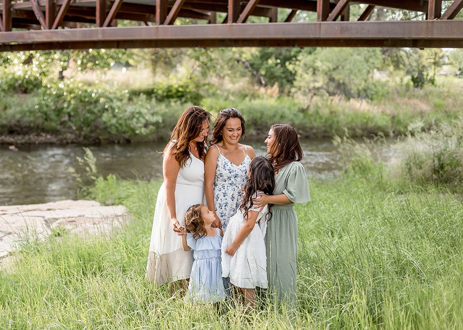 A mother and three daughters in floral and pastel dresses pose together in a lush green field near a creek.