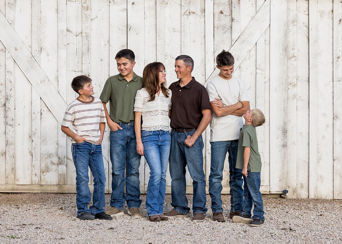 Family of five smiling and laughing together against a rustic white barn wall, wearing casual jeans and tops.