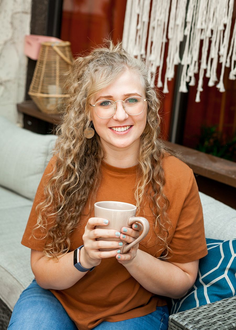Smiling woman with curly blonde hair holding a beige mug, wearing an orange t-shirt, sitting on a cozy outdoor patio.