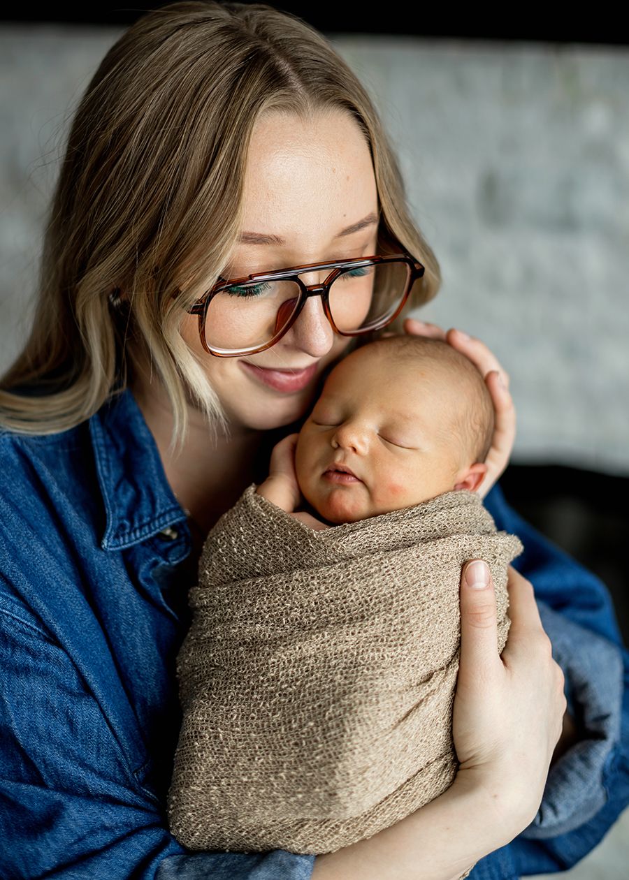 A young woman with glasses tenderly holds a sleeping newborn wrapped in a cozy brown swaddle blanket.