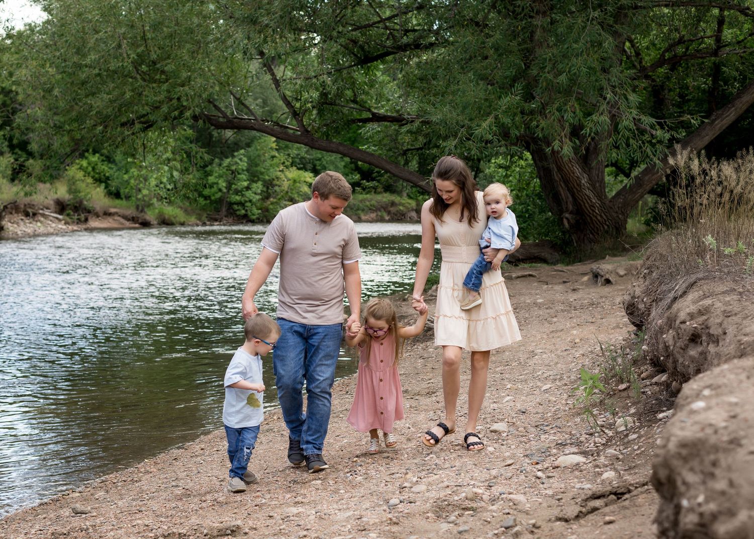 Family of five strolling along a scenic riverbank with green trees in the background.