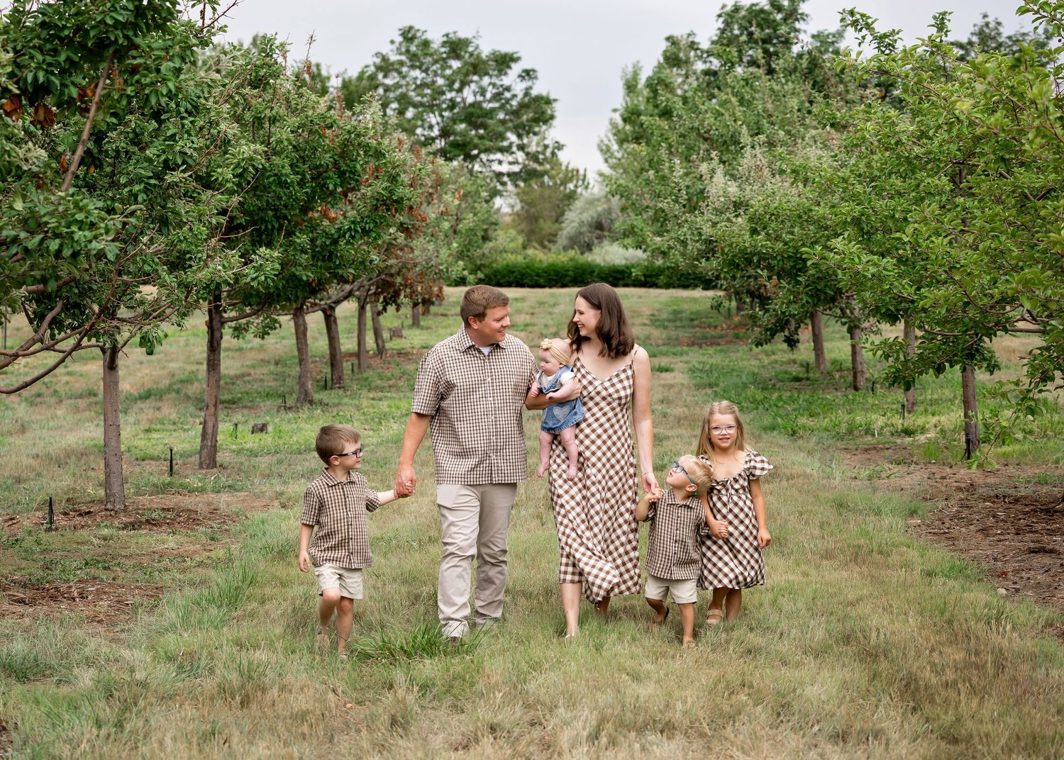 Family of five walking through a lush green orchard, smiling and holding hands on a warm summer day.