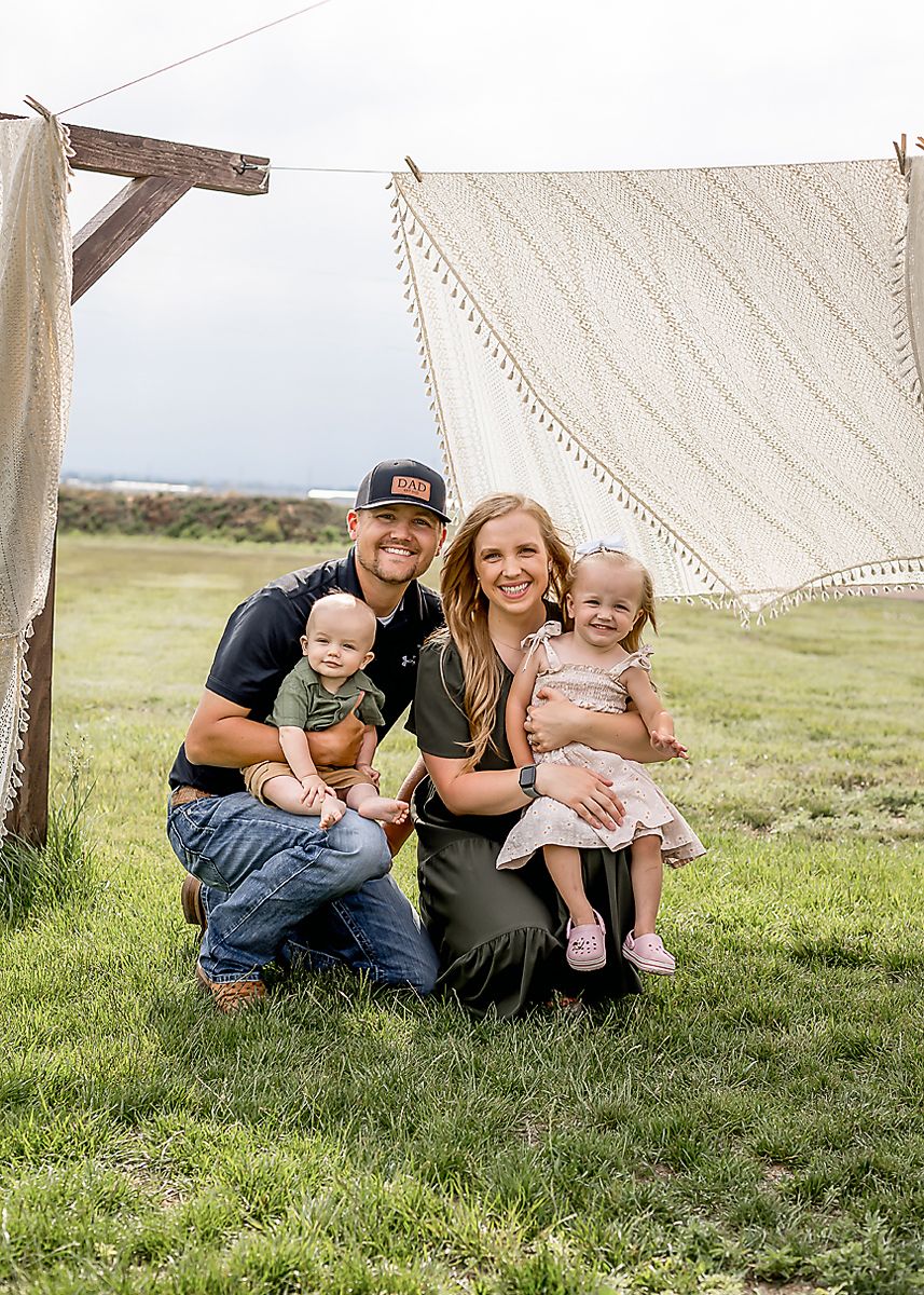 Young family with two toddlers kneeling under a macrame backdrop outdoors.
