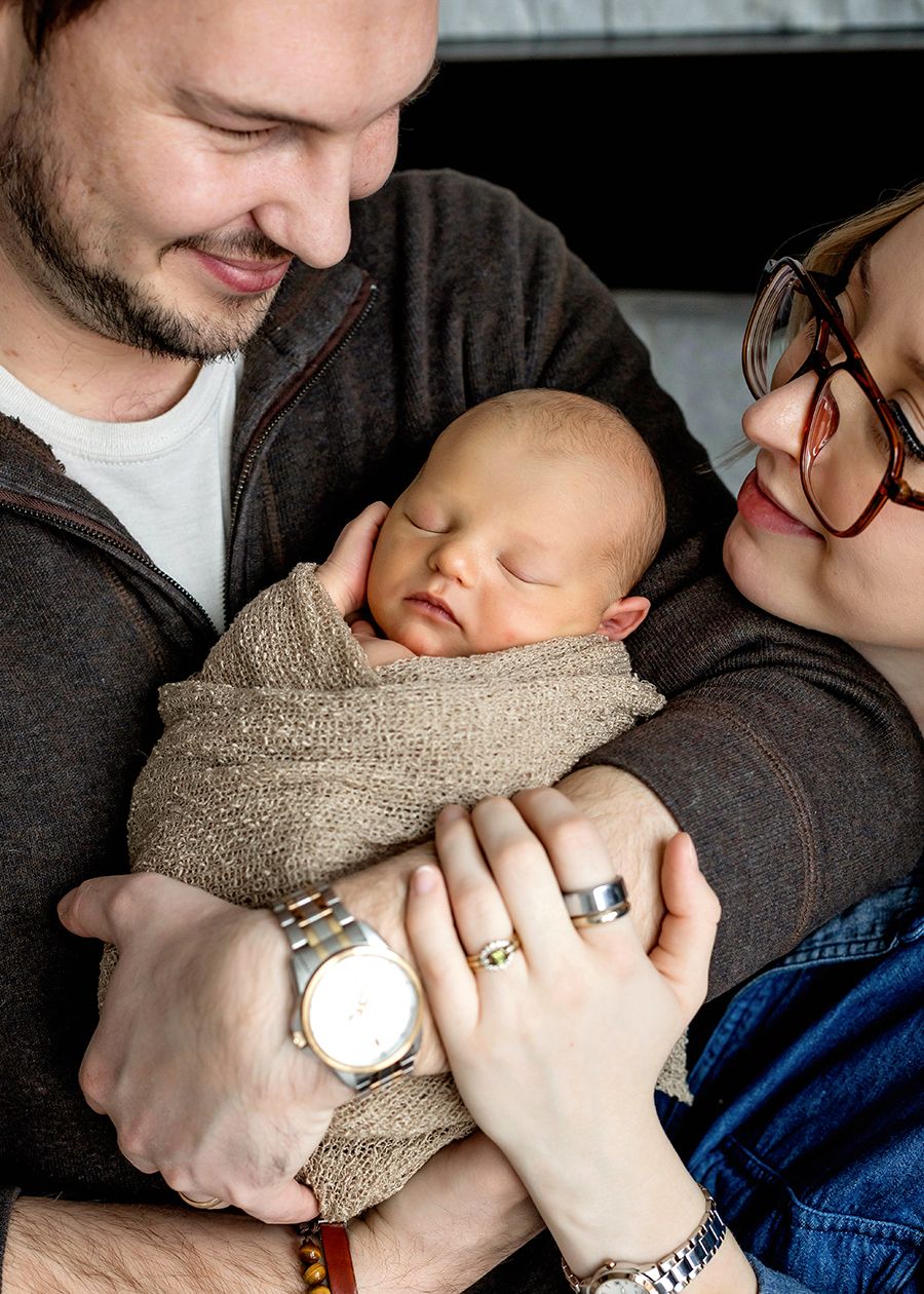 Parents lovingly cradle their sleeping newborn wrapped in a tan swaddle, mom wearing red glasses, dad wearing a watch.