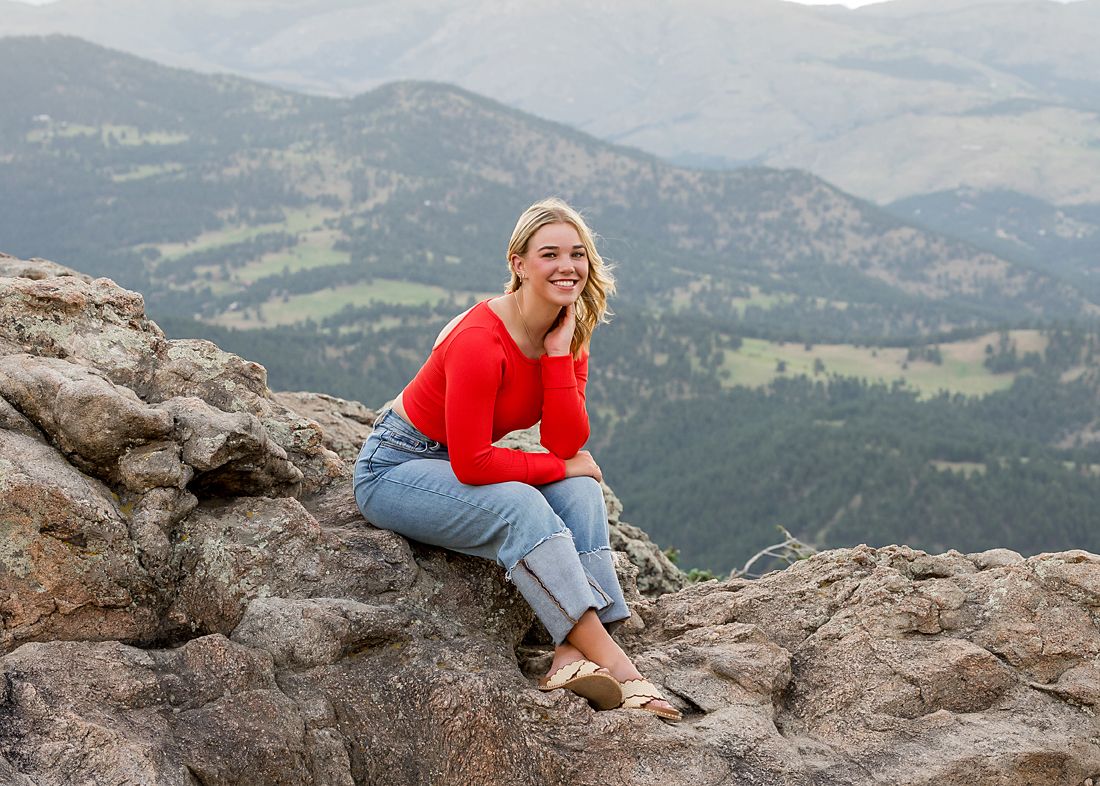 Smiling blonde teen girl sits on rocky mountain summit with scenic valley views behind her.