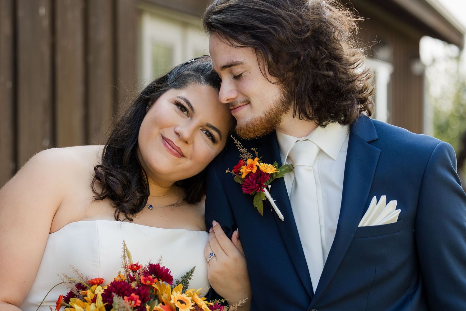 A couple in wedding attire embrace while holding autumn-colored bouquets during an outdoor ceremony.