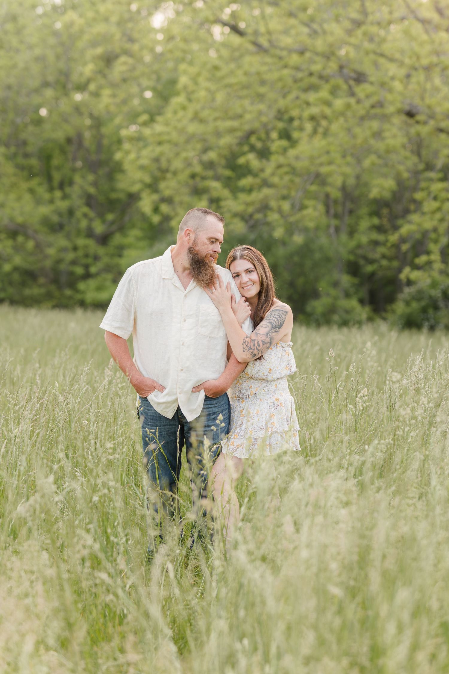 A couple shares a romantic moment in a sunlit meadow with tall grass and green trees in the background.