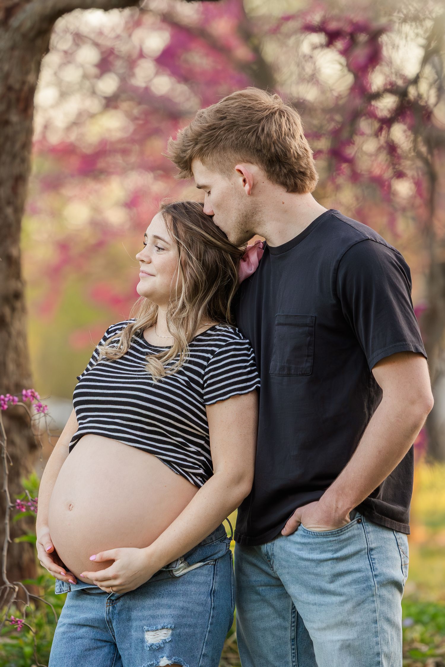 A couple shares tender moments in a pink blossom-filled garden during a spring maternity photoshoot.