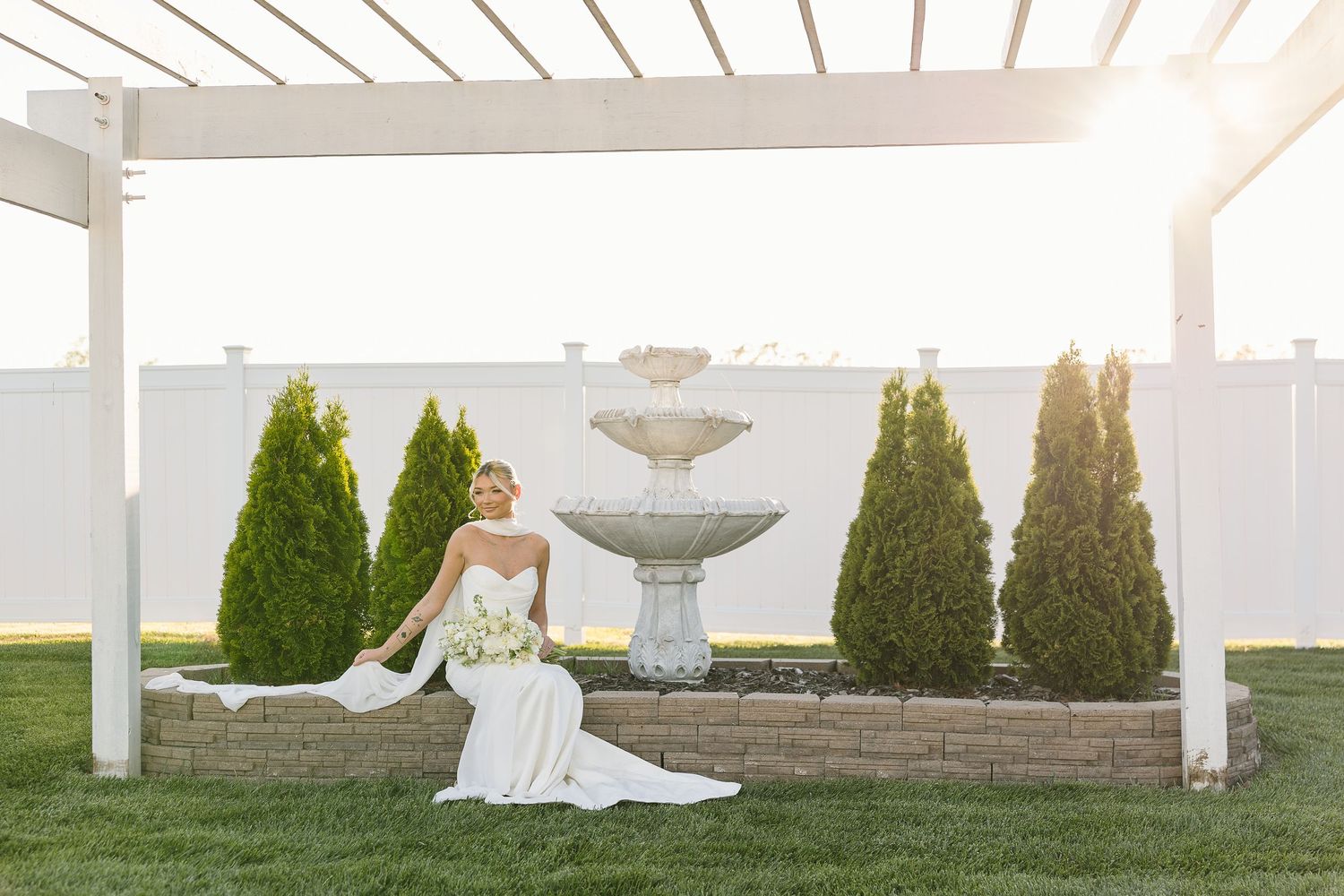 An elegant bride in a white strapless gown sits gracefully beside a tiered fountain with evergreen shrubs in the background.